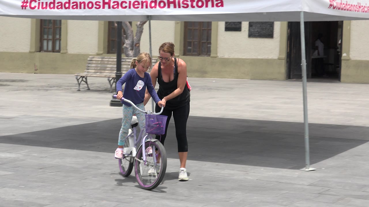 Woman steadies a girl on a purple bicycle in a plaza.