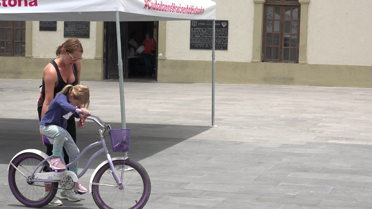 In Los Cabos, Mexico, an adult woman stands behind a young girl seated on a small purple bicycle in bright light on a paved plaza.