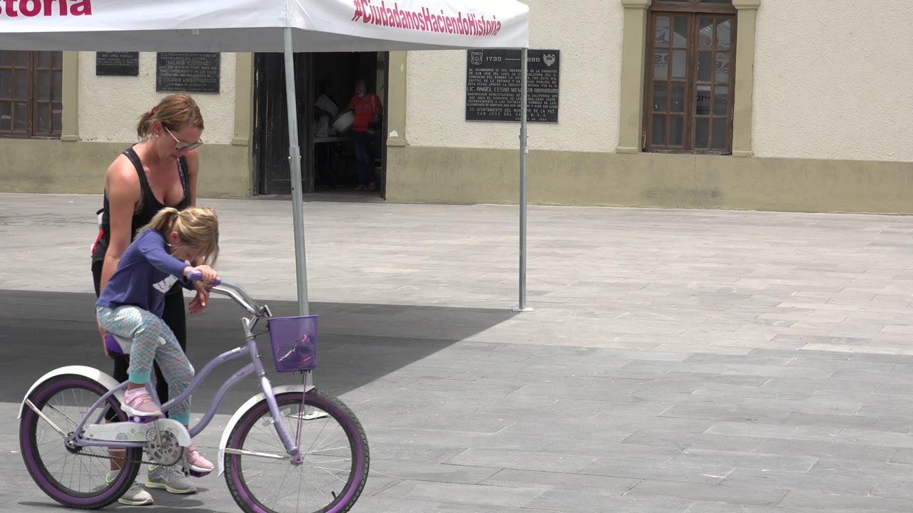 The child leans forward over the handlebars of the purple bicycle while the adult remains directly behind her on the Los Cabos plaza.