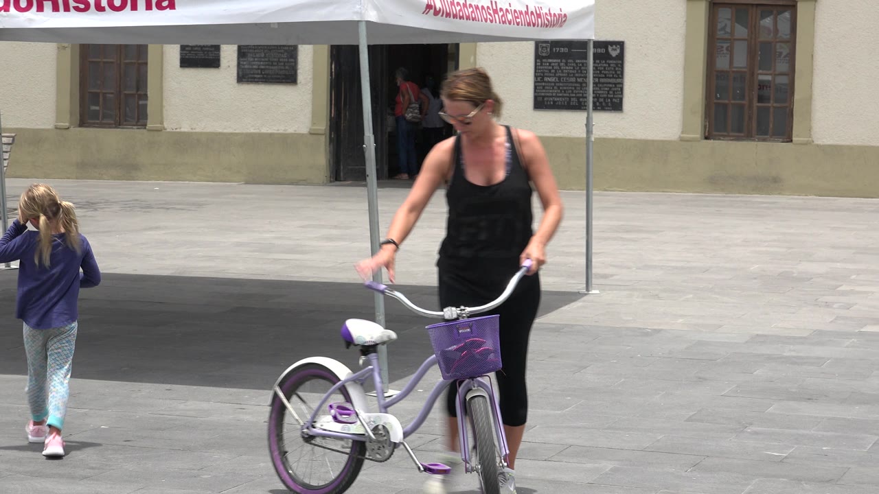 In Los Cabos, Mexico, the woman stands closer to the purple bicycle as the young girl moves farther into the open paved space under the edge of the canopy shade.