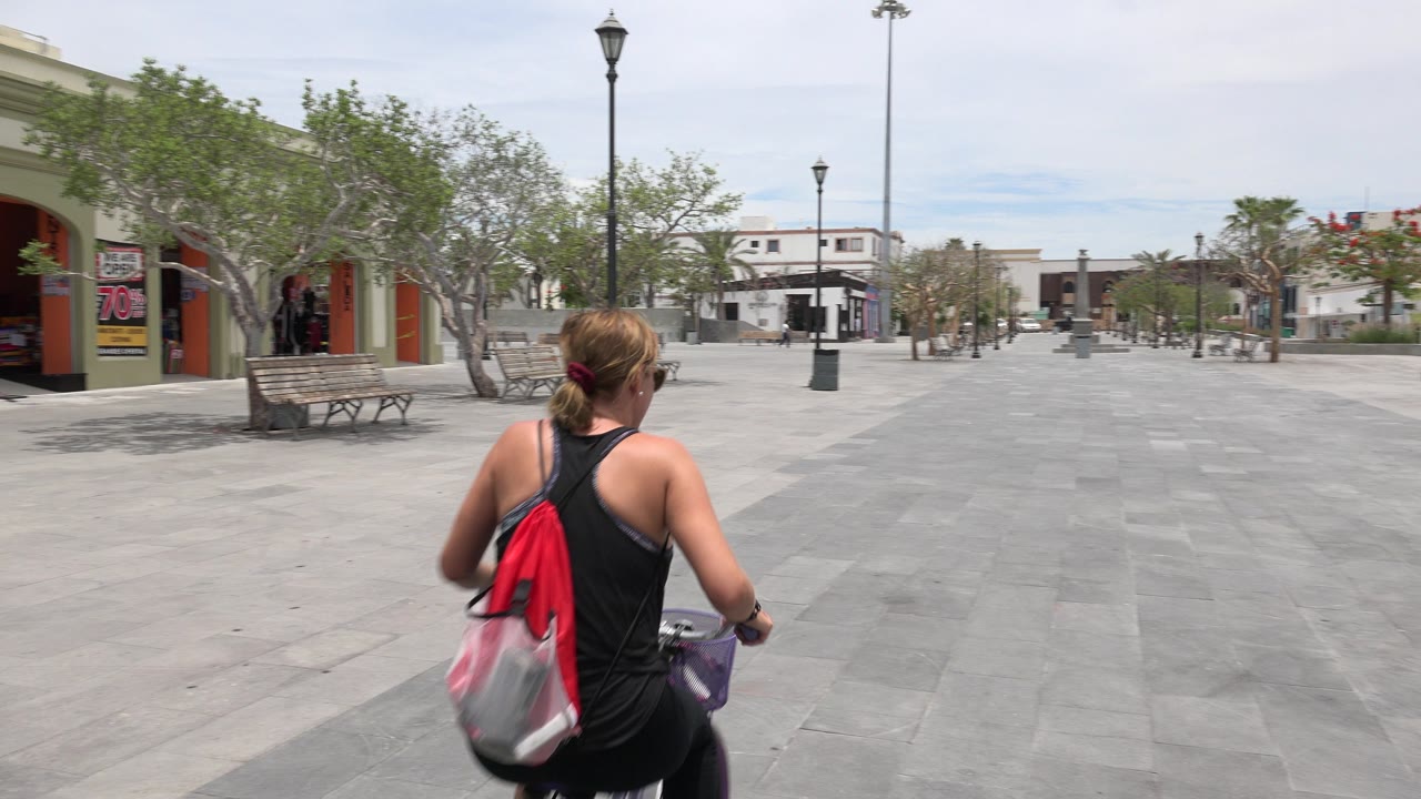 The cyclist continues away along the gray paved walkway in Los Cabos, Mexico, with plaza benches and trees lining both sides of the frame.