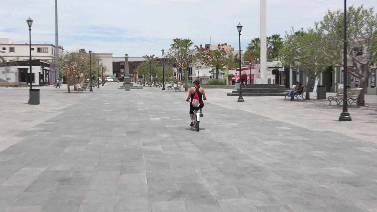 A woman rides a small bicycle away from the camera across a broad stone plaza in Los Cabos, Mexico, with lamp posts, benches, trees, and storefronts ahead.