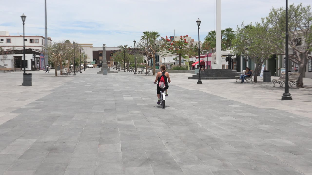 The bicyclist remains centered in the Los Cabos plaza, moving away across gray paving toward lamp posts, benches, small trees, and low-rise buildings.