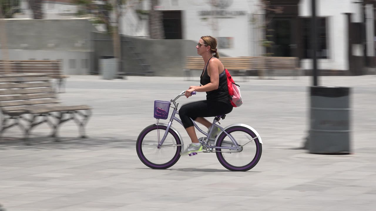 The cyclist moves farther across the Los Cabos plaza, passing wooden benches, a dark trash can, and pale paving under strong daylight.