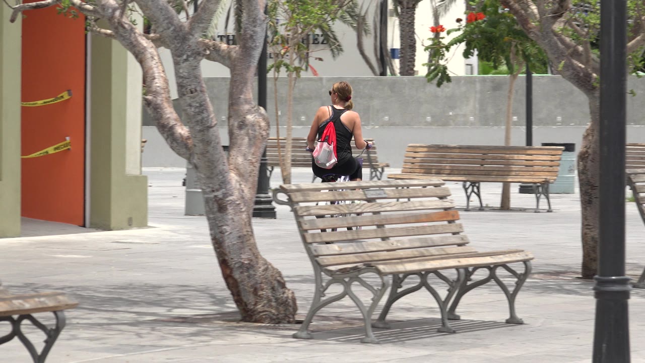 A woman rides a bicycle in a Los Cabos, Mexico park, with wooden benches, tree trunks, pale pavement, and an orange wall detail visible behind her.