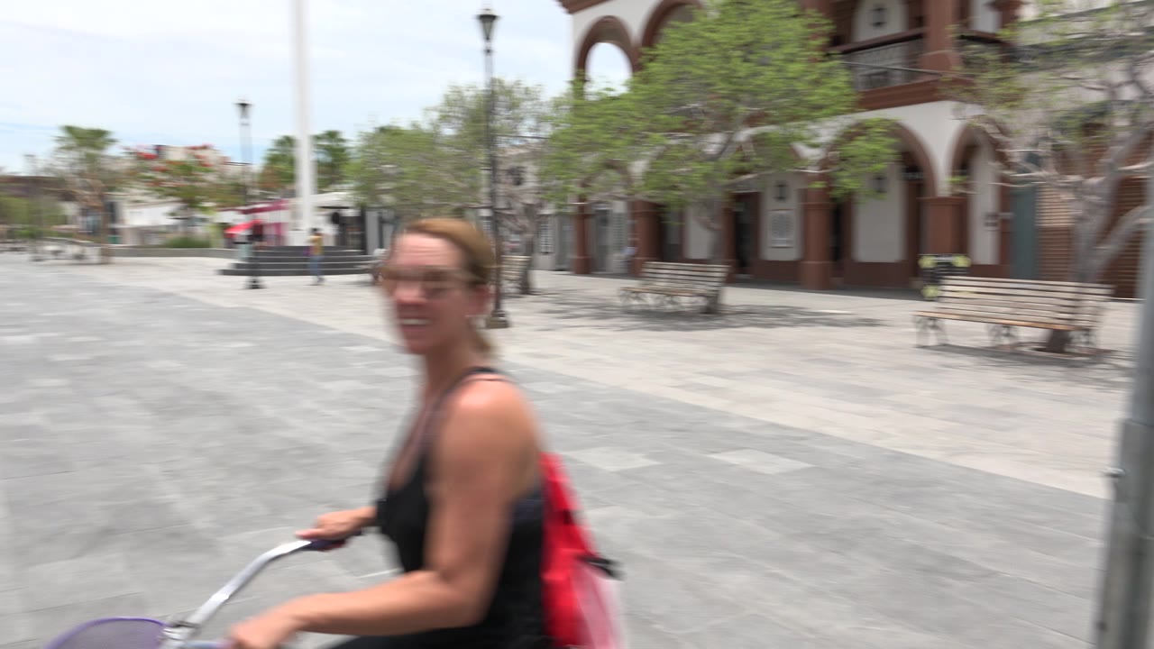Woman on bicycle passing close to camera in a paved plaza.