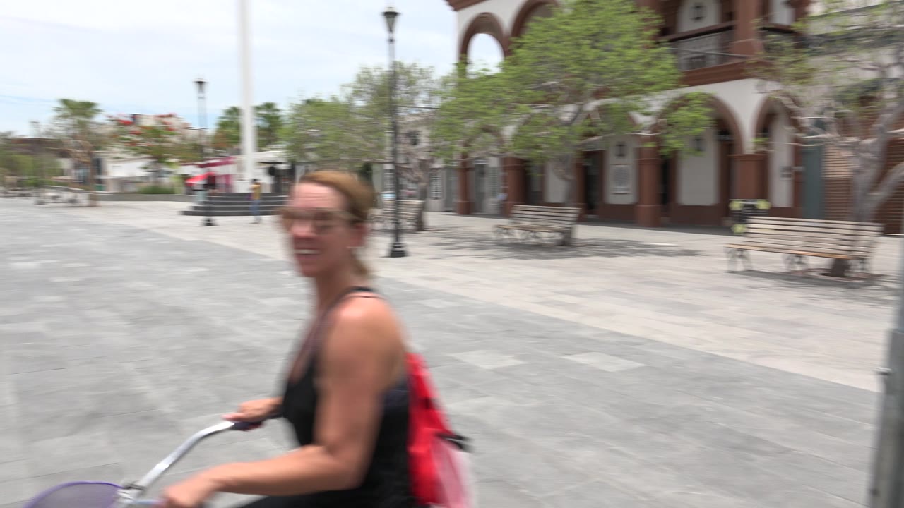 The woman rides near the camera in the Los Cabos plaza, with motion blur on her face and shoulders, a red backpack, gray paving, benches, trees, and lamp posts visible behind her.
