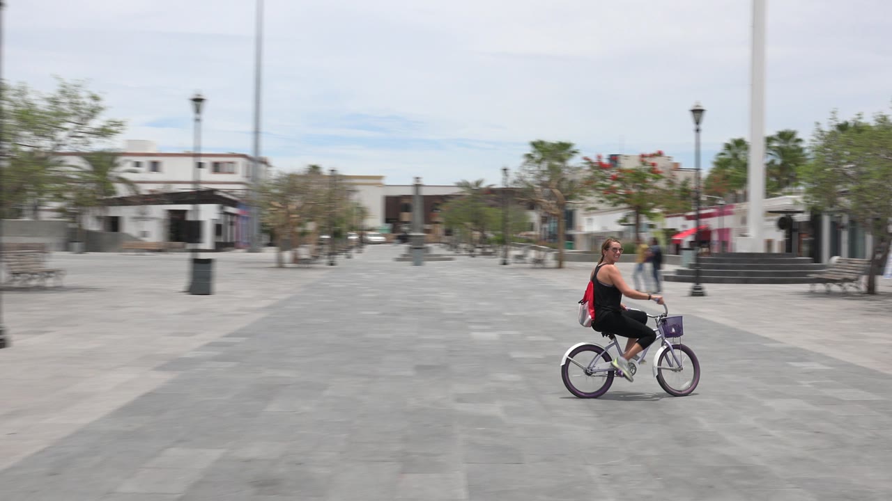 The adult on the purple bicycle continues across the Los Cabos plaza, framed by wide gray paving, lamp posts, benches, trees, and small figures walking in the background.