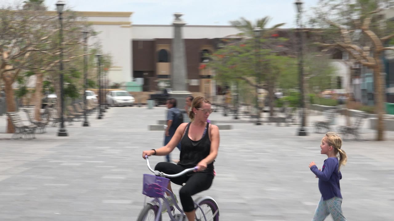 In Los Cabos, Mexico, a woman rides a purple bicycle at the left of the frame while a young girl runs in the midground across a paved plaza with benches and lamp posts.