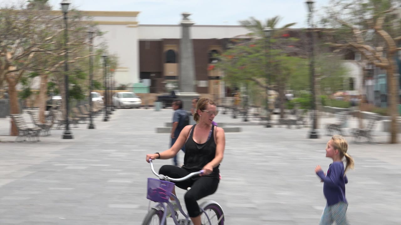 The woman continues cycling through the Los Cabos plaza as the young girl runs from the right side of the frame, with benches, trees, lamp posts, and parked vehicles behind them.