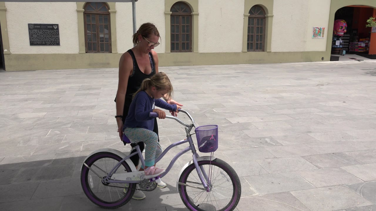 Woman stands beside a girl seated on a small bicycle in a paved square.