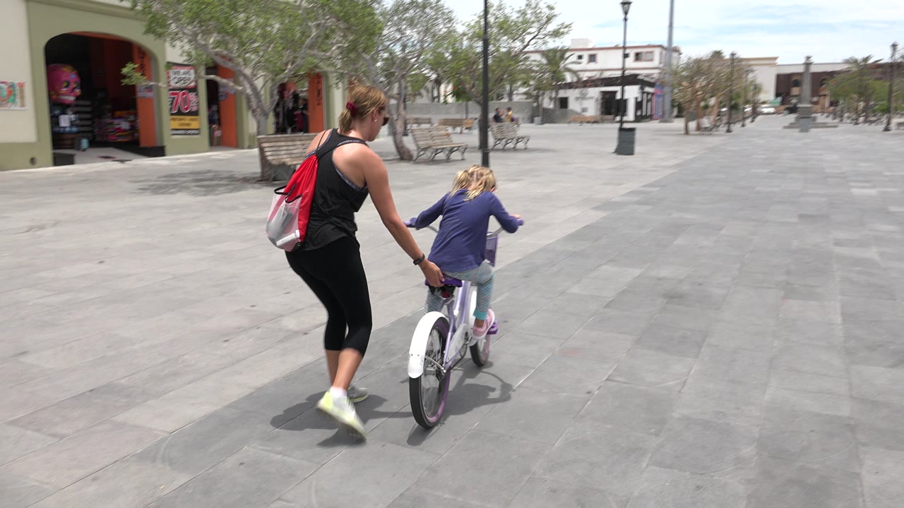 Woman helping a young girl ride a small bicycle in an open plaza.