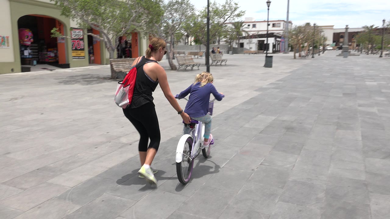 The mother and child continue forward through the Los Cabos plaza, with the woman holding near the bicycle seat and a sale sign visible by the left storefront.