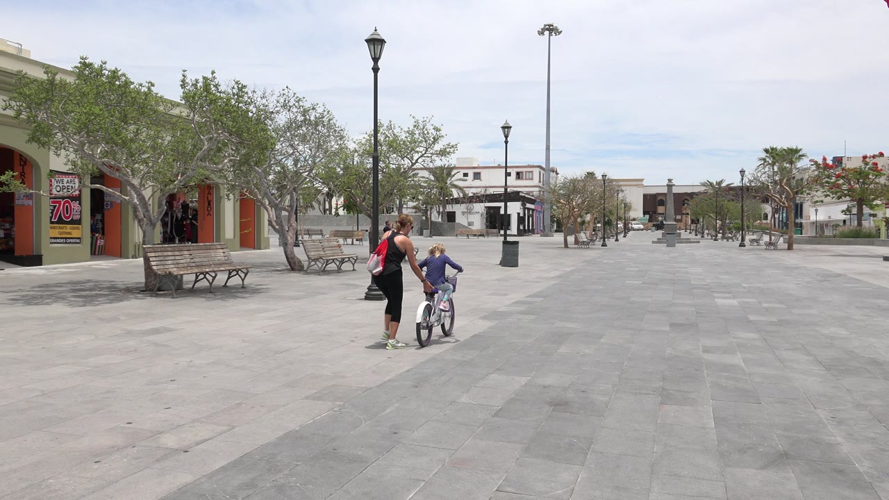 Woman helping a child on a small bicycle in a paved plaza.