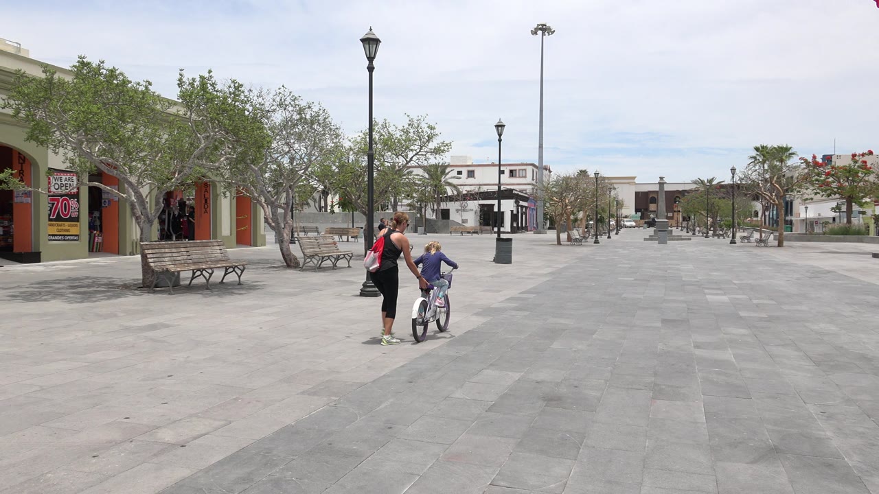 One second in, the woman keeps close to the child on the bicycle as they move across the gray paving, with lamp posts and shaded storefronts in the background.