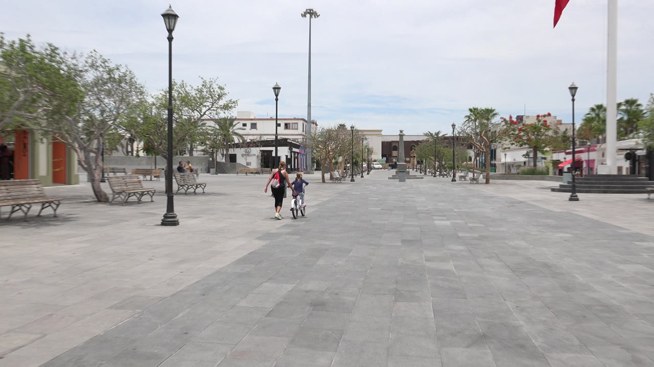 The mother stays beside the child on the bicycle as they move across the open Los Cabos plaza, with benches, trees, lampposts, and white buildings behind them.