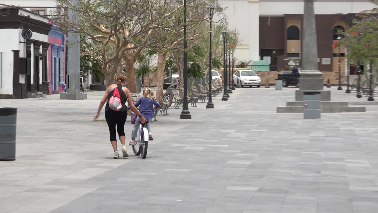 In Los Cabos, Mexico, a woman with a backpack walks behind a young child on a small bicycle across a wide paved plaza with benches, trees, and buildings ahead.