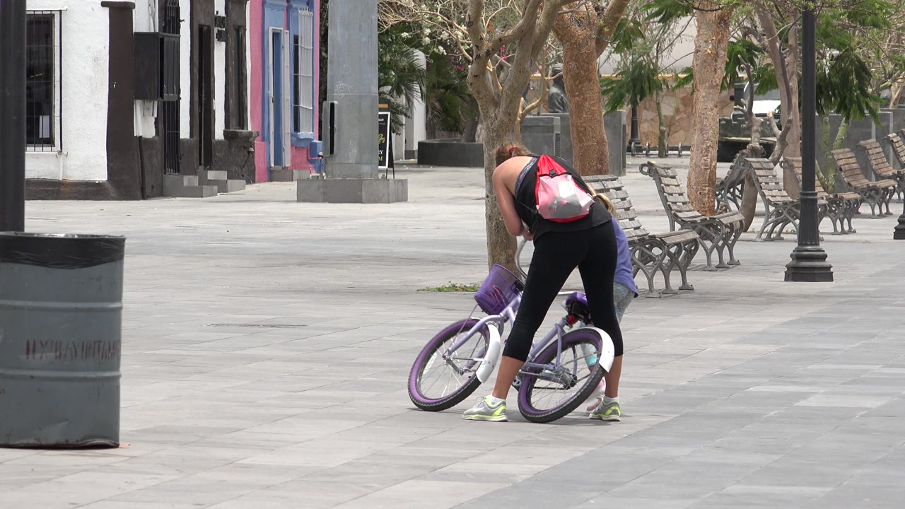 At the start of the Los Cabos clip, a woman with a red backpack bends over a purple and white bicycle on a broad paved plaza with benches and trees behind her.