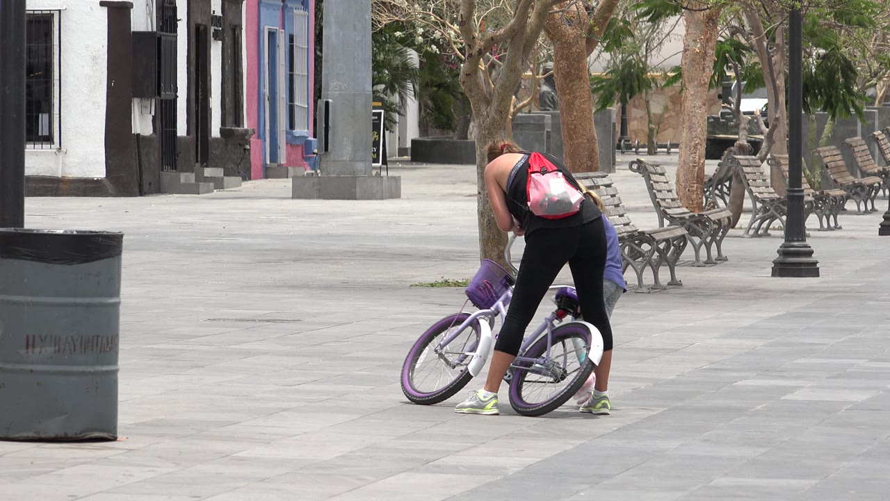 One second in, the woman remains bent over the bicycle in the Los Cabos plaza, with empty paving, benches, lamp posts, and tree trunks filling the background.