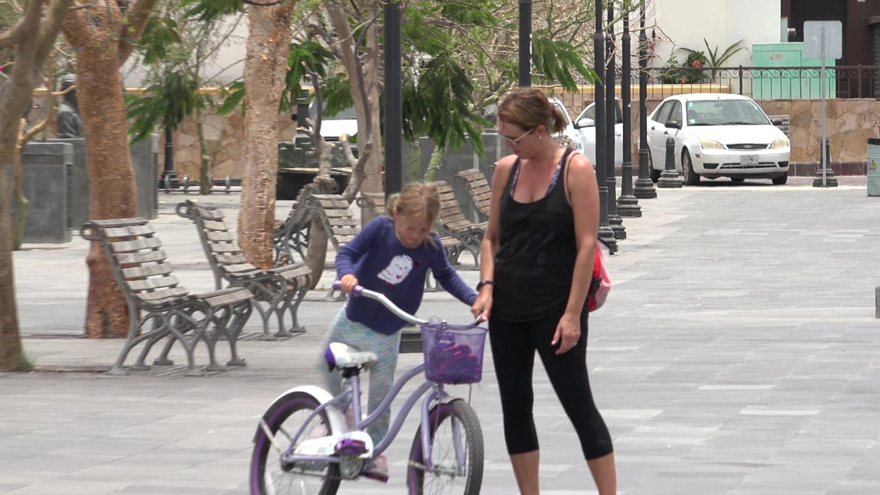 Mother walks beside a girl holding a purple bicycle.