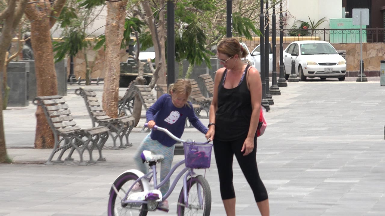 The girl leans toward the purple bicycle while the adult woman walks beside her on the paved Los Cabos walkway, with benches and a white car behind them.