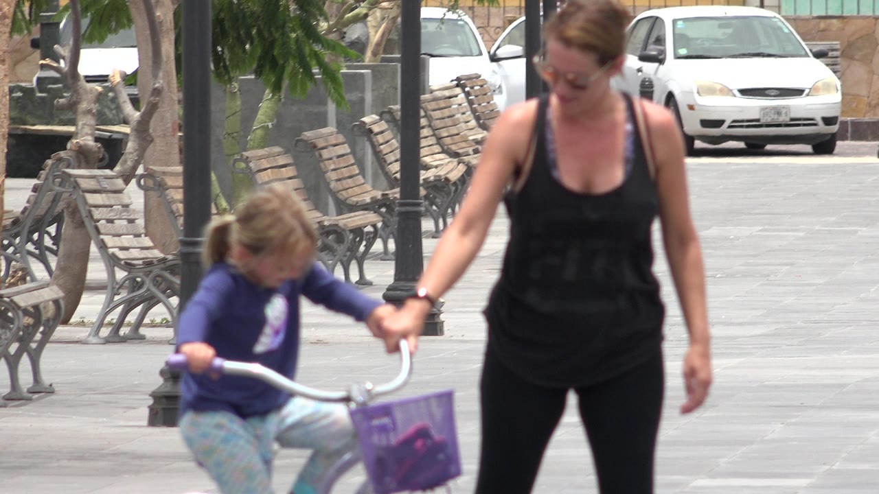 Woman walking beside a child on a purple bicycle in a paved plaza.
