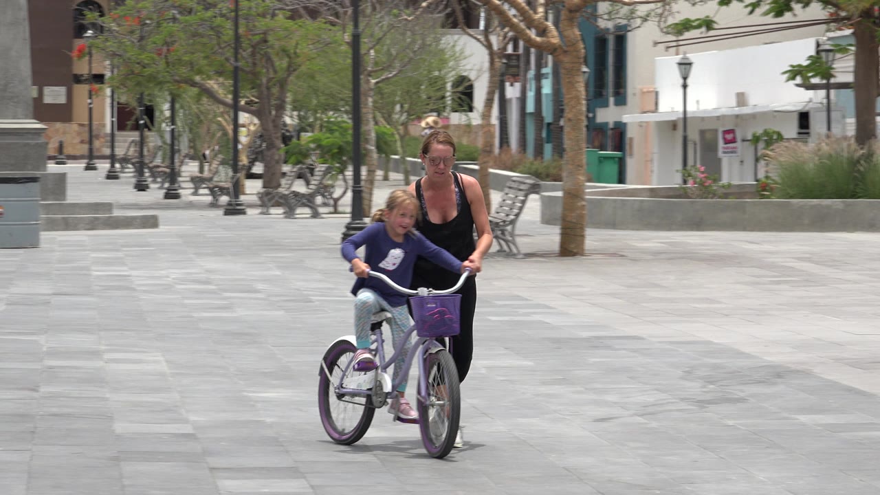 In Los Cabos, Mexico, a woman walks behind and beside a young girl riding a small bicycle across a broad paved plaza with benches and trees behind them.