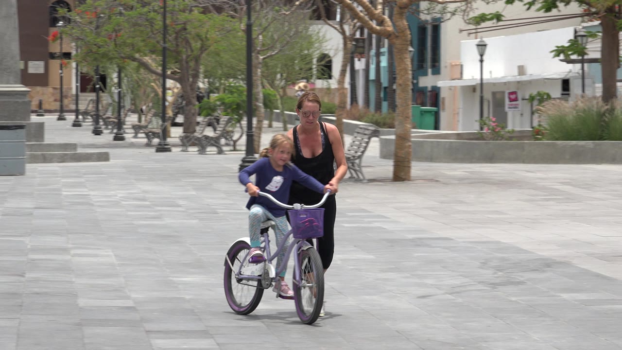 The girl continues toward the camera on her bicycle in Los Cabos, Mexico, with the woman slightly behind her and rows of benches and trees lining the plaza.
