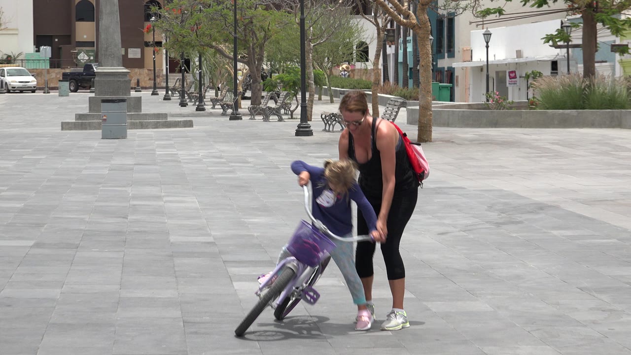 Woman steadies a young girl leaning on a purple bicycle in a paved plaza.