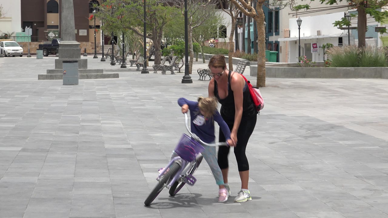 In Los Cabos, Mexico, the child and bicycle remain angled while the woman braces the handlebars and seat area on the paved square.