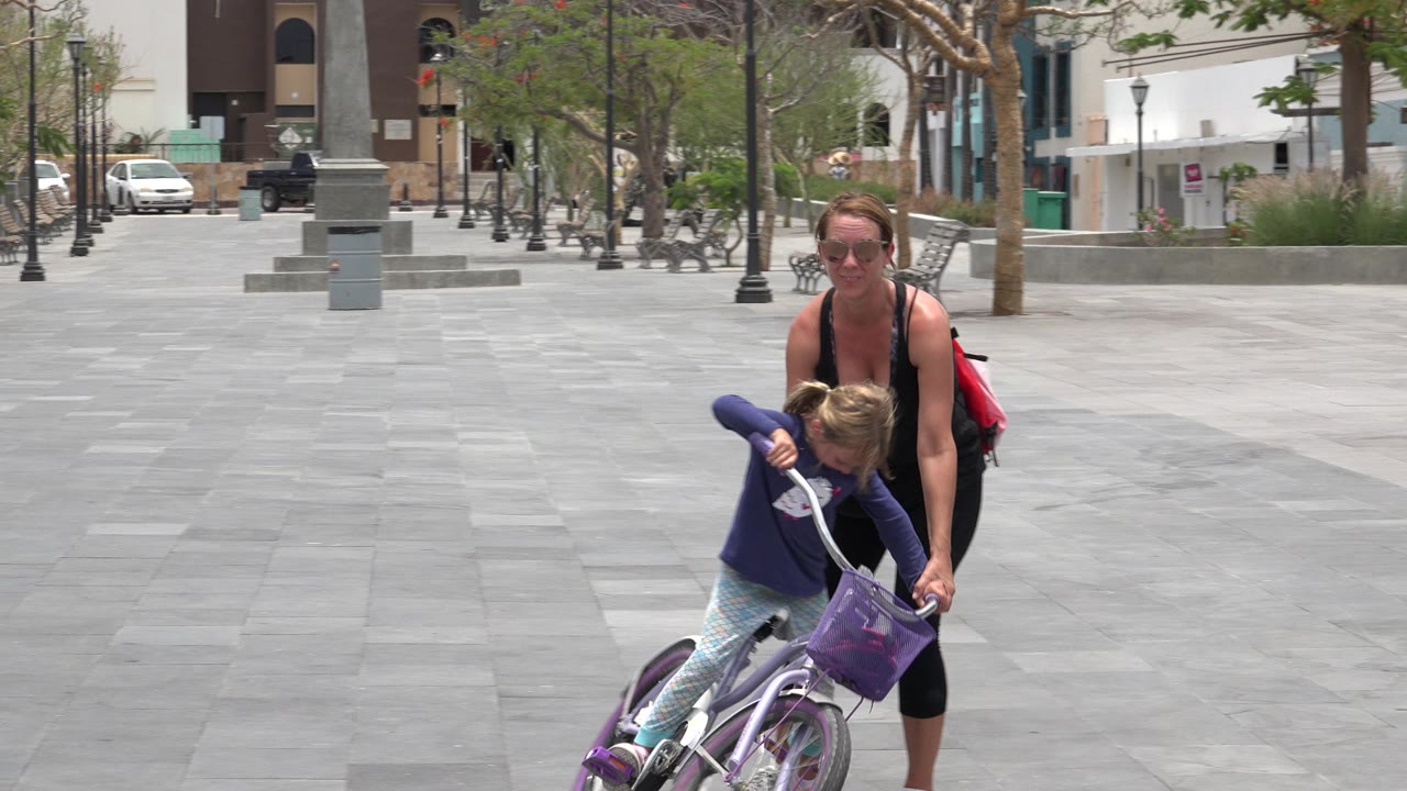 Woman helping a young girl on a purple bicycle in a paved plaza.