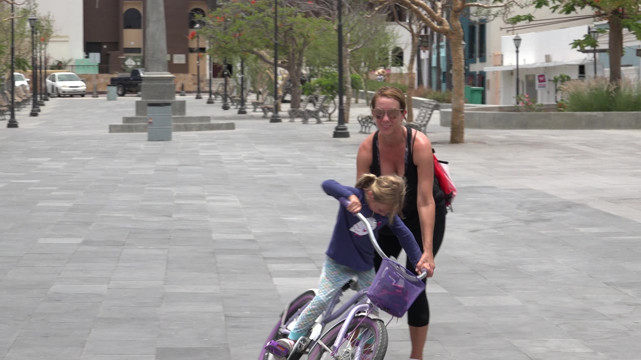 The girl leans forward over the purple bike while the woman follows closely in the open Los Cabos plaza, with trees and buildings behind them.
