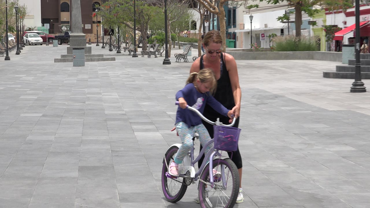 The girl pedals the small bicycle forward in Los Cabos while the woman stays beside and slightly behind her on the open paved plaza.