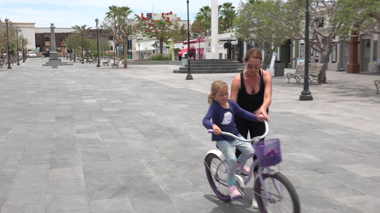 In Los Cabos, Mexico, a woman steadies a young girl on a white and purple bicycle near the foreground of a wide paved plaza with benches and palm trees behind them.