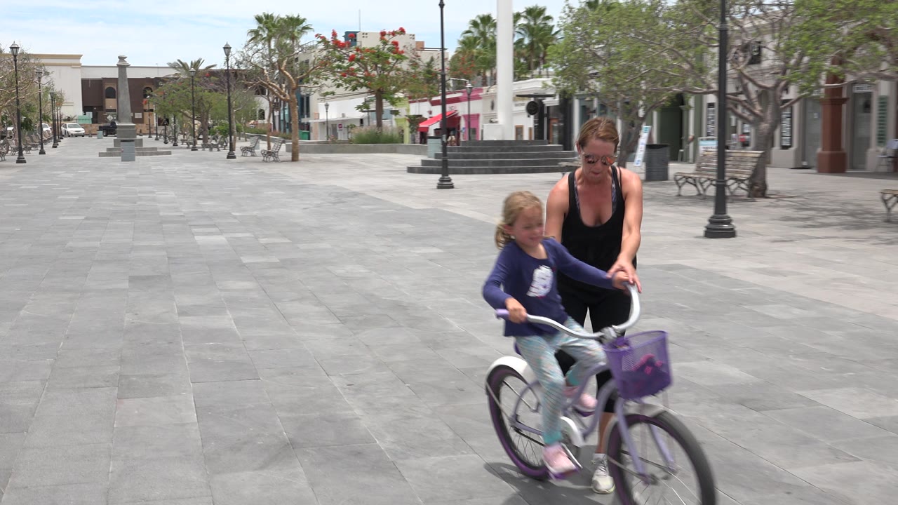 In Los Cabos, Mexico, the child pedals the small bicycle while the woman walks beside her, with gray paving, trees, lamps, and shaded storefronts in the background.