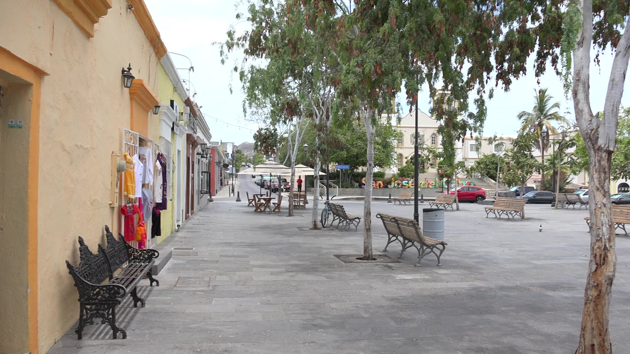 Empty paved square with benches, trees, storefronts, and parked cars.