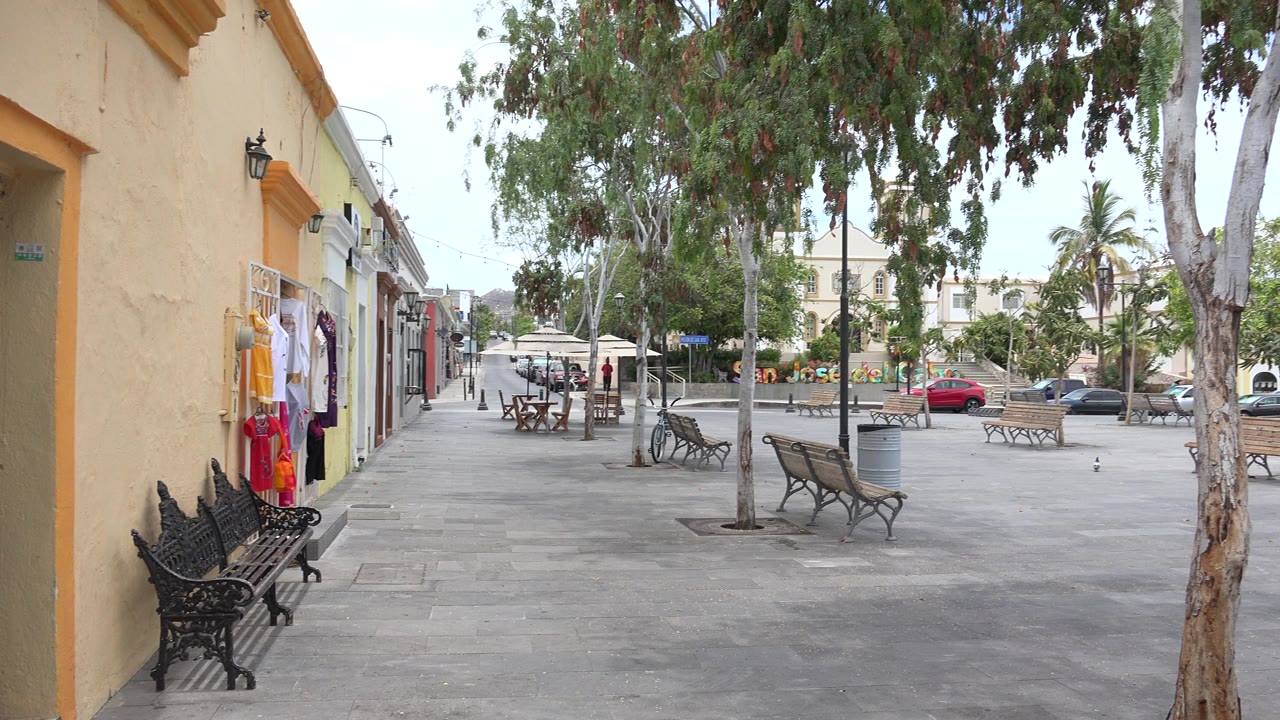Second frame of the Los Cabos square shows the same empty walkway, shaded benches on the left, trees across the pavement, trash cans, parked cars, and no visible people.