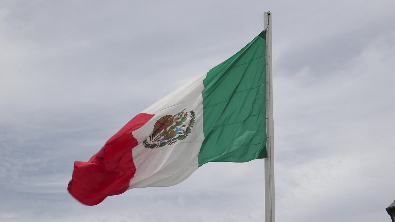 Mexican flag waving beside a pole under gray clouds.