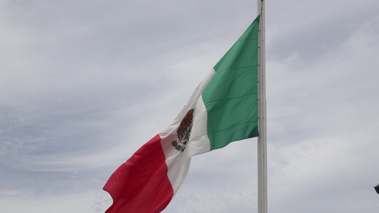 Mexican flag partly unfurled beside a flagpole under gray clouds.