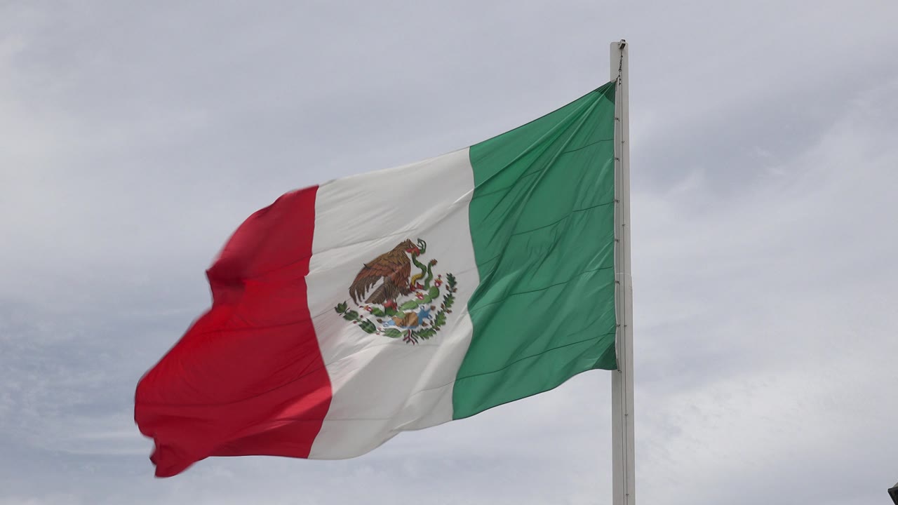 Mexican flag in Los Cabos, Mexico spreads left from a white pole under a gray cloudy sky, with no visible people.