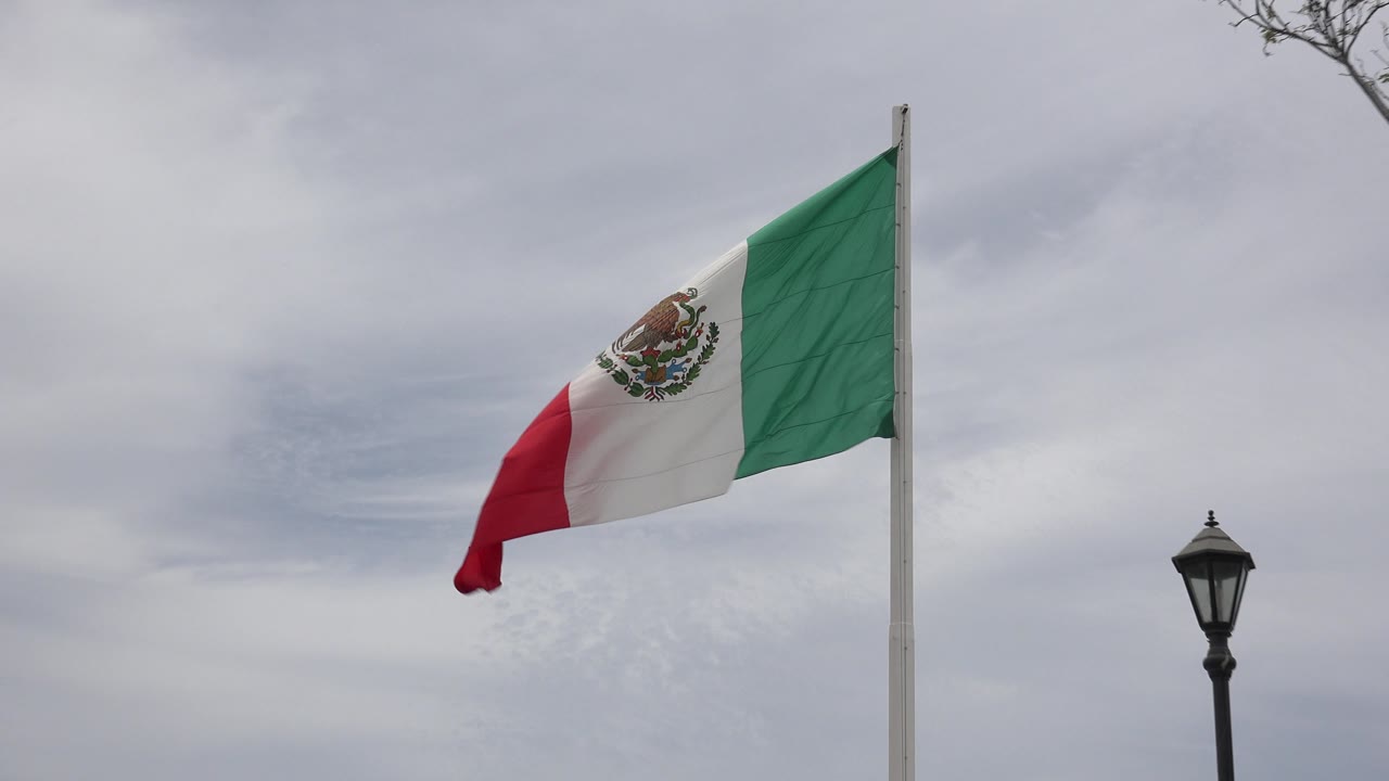 Mexican flag lifting above a lamp post against clouds.