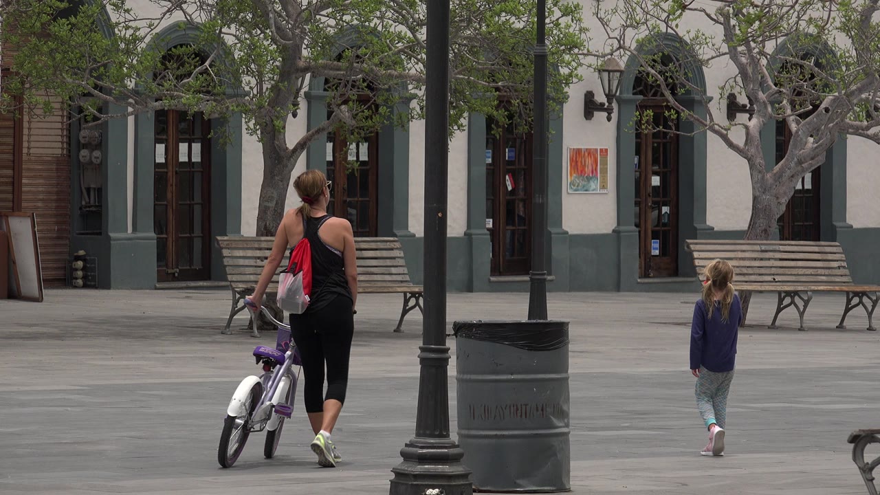 Adult walking a bicycle while a child walks ahead in a paved plaza.