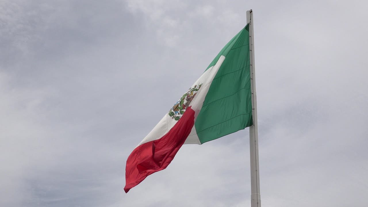 Mexican flag in Los Cabos, Mexico, partly wrapped and folding near the pole against soft gray clouds, with no visible people.