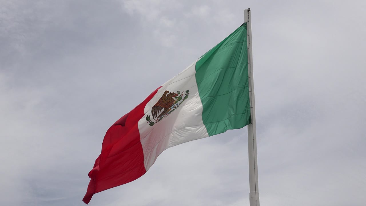 Mexican flag in Los Cabos bends left from a flagpole beneath a gray overcast sky, with no visible people.