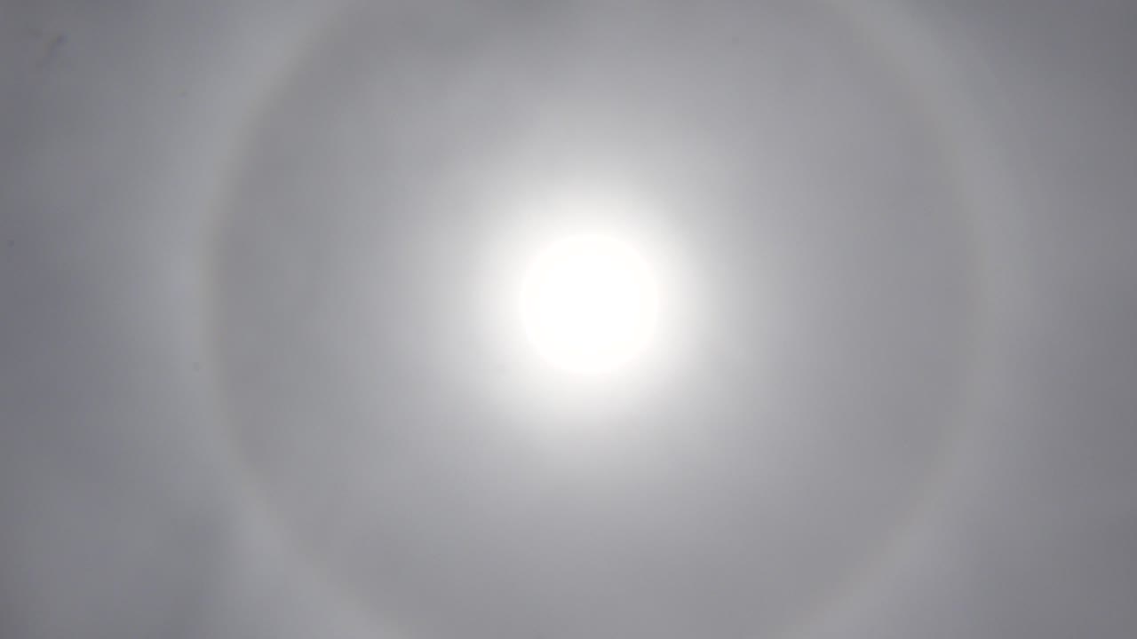 White sun holds near the center of the Los Cabos sky, with gray haze and a pale rainbow halo, no visible people.