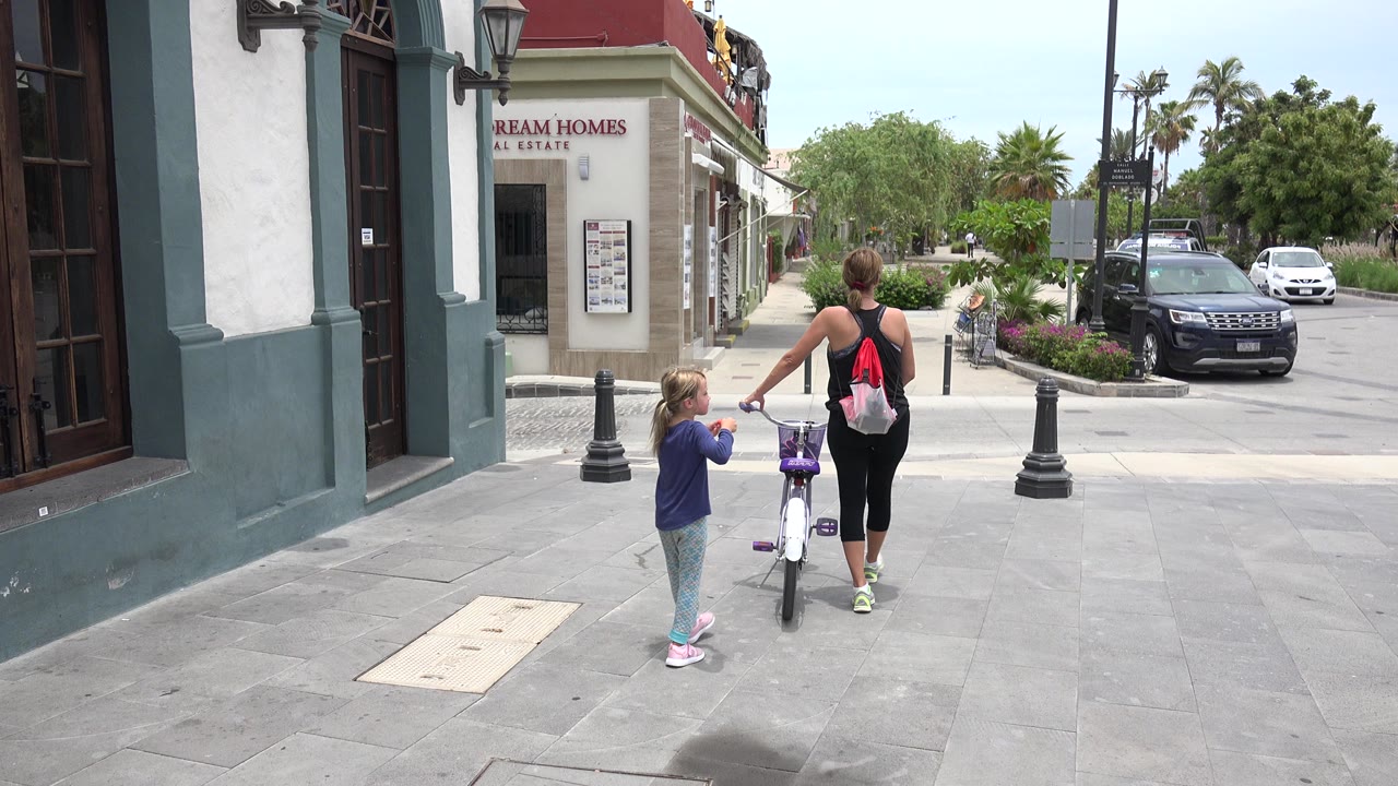 Woman pushes a child bicycle while a young child follows on a sidewalk.