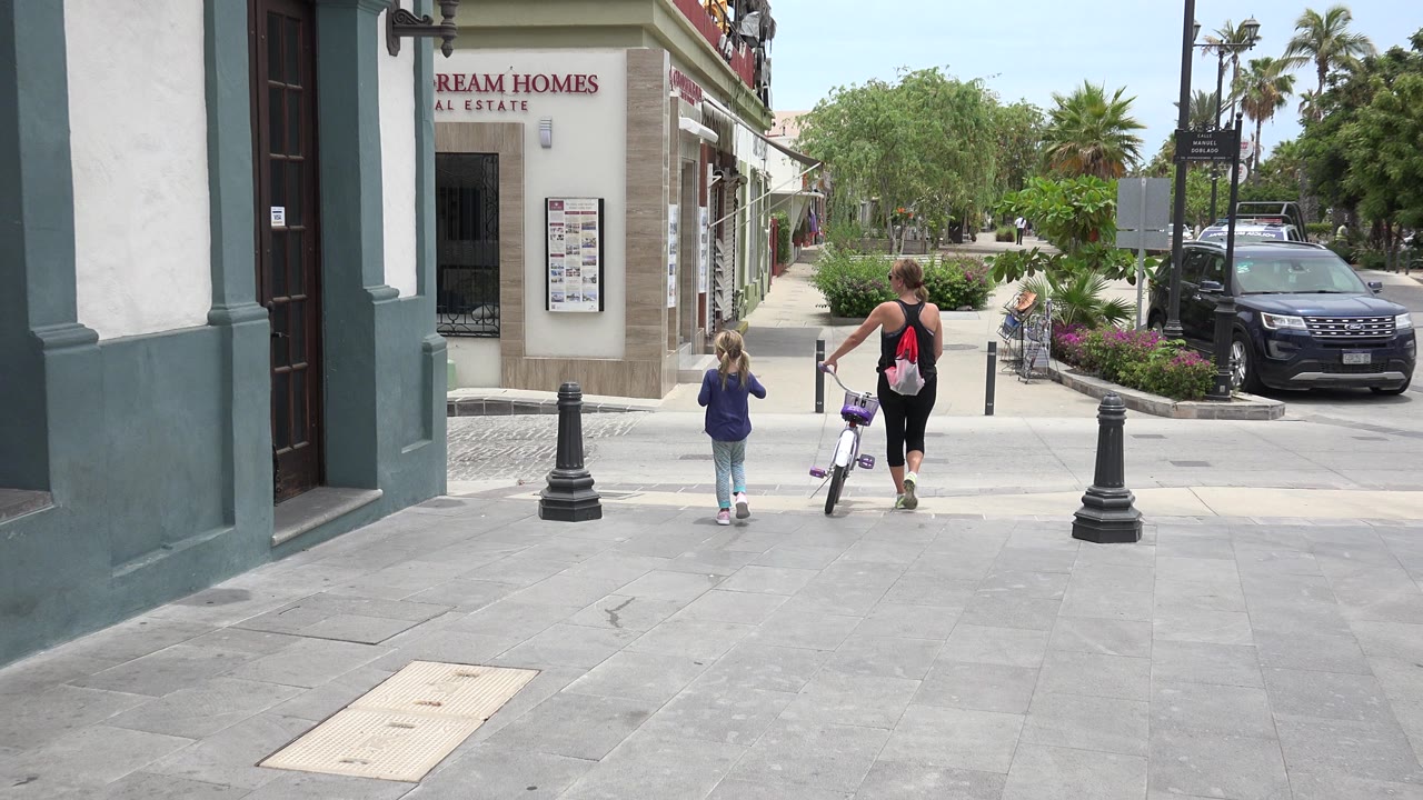 The woman walks the small bicycle farther along the Los Cabos sidewalk while the child stays to the left, framed by bollards, a real estate storefront sign, shrubs, and a parked SUV.
