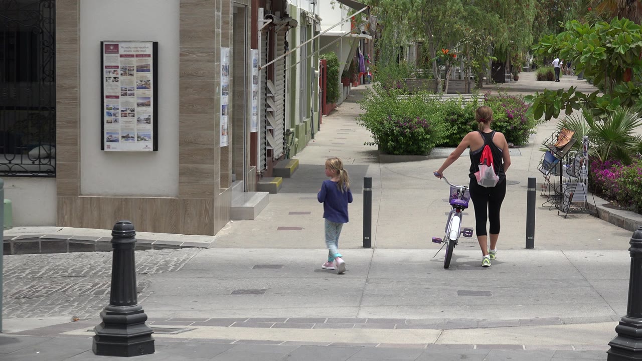 The child and the adult with the bicycle are farther inside the Los Cabos pedestrian area, with storefront columns on the left and leafy shrubs and benches on the right.