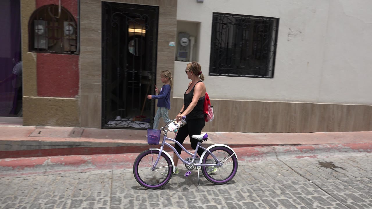 Woman with bicycle and girl walking past barred windows on a cobblestone street.