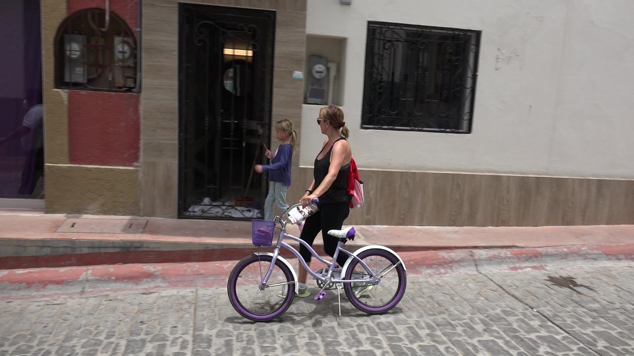 In Los Cabos, Mexico, the woman and bicycle continue in profile as the girl walks ahead near a black metal doorway and barred window.